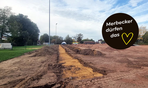 ehemaliger Ascheplatz / Baufläche<br />
Blick über die große Baustelle des neuen Sportplatzes des SV Merbeck: Auf dem ehemaligen Sportplatz sind Erdarbeiten und mehrere aufgeschüttete Erdhügel zu sehen. Eine Einblendung mit gelbem Herz und dem Schriftzug „Danke“ begleitet das Foto.